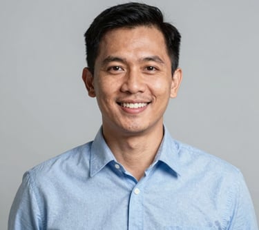 A professional corporate headshot of a friendly Southeast Asian / Indonesian man in his 30s wearing a light blue shirt. He has a warm smile and is positioned against a simple, light gray studio background. The style is modern, sharp, and clean.