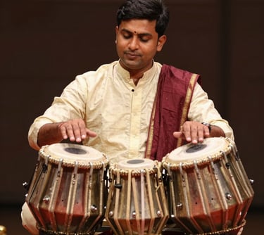 A professional South Asian / Indian male musician playing a set of traditional tabla drums, focused expression, wearing ethnic attire in Soft Cream and Deep Burgundy colors.