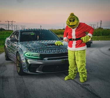 The Grinch in a Santa suit stands next to a gray Dodge Charger wrapped in festive Christmas lights at sunset.