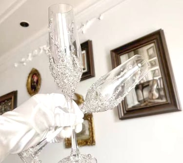 A gloved hand holds luxury crystal champagne flutes decorated with silver glitter and rhinestones.