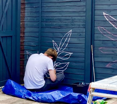 Muralist sits on floor painting graphic plant on grey wooden garden fence.