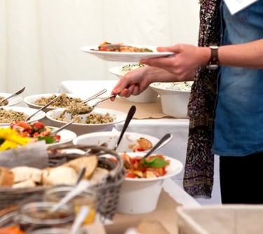 A person serving fresh healthy food from a catered buffet line featuring quinoa salad and bread.