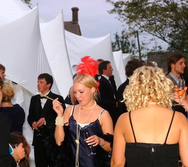 Guests at a Great Gatsby themed party in 1920s flapper costumes and formal suits outside a white tent.
