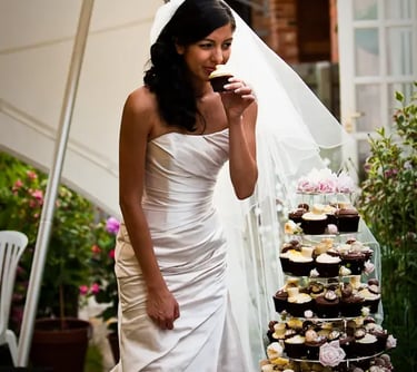 A bride in a white wedding gown eating a chocolate cupcake by a tiered cupcake tower.