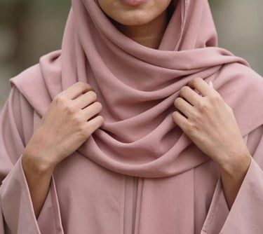 A close-up photograph of a woman wearing a dusty rose abaya, her hands gently adjusting the drape of her hijab, captured in natural, soft lighting. Middle Eastern / Gulf setting.