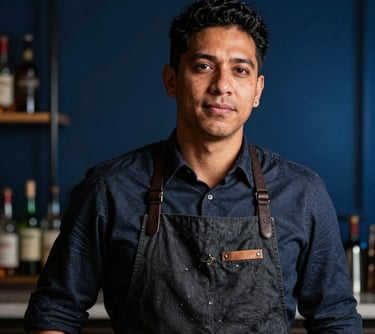 Professional portrait of a master mixologist in a smart dark apron, South American / Brazilian, focused and refined, with a backdrop of soft midnight blue lighting in a high-end bar setting.