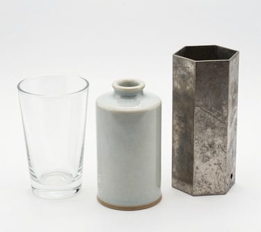 A sleek, modern vase with a smooth matte finish sitting on a wooden shelf against a soft neutral wall.