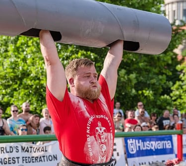A strongman competitor in a red shirt performs a heavy log press lift at an outdoor competition.