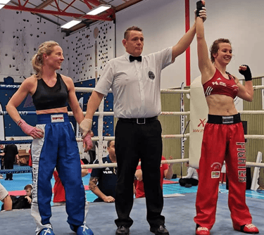 Referee raising the hand of a female kickboxer in victory inside a competitive boxing ring.