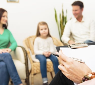 A woman sitting in a chair, holding a clipboard, surrounded by her family. Image relate to autism and ADHD in Vadodara