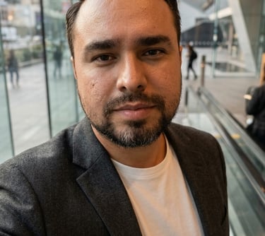 Professional man in a gray blazer and white t-shirt standing in a modern office building with city views.