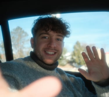 Smiling young man with curly hair waving while sitting in the back seat of a car.