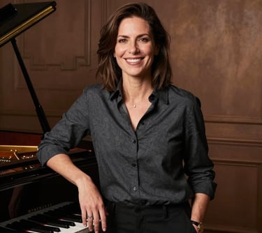 A professional portrait of a smiling woman in a dark charcoal shirt, standing next to a grand piano in a studio with rich bronze lighting.