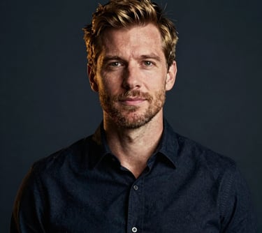 A professional portrait of a man in a modern studio setting. He is wearing a dark navy shirt. The lighting is sophisticated, using burnished gold tones to create depth against a deep charcoal navy background.