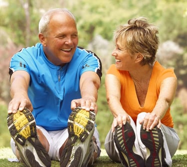 Un homme et une femme souriant, assis sur l’herbe, dans un mouvement d'assouplissement main-pied.