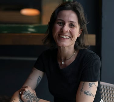 Smiling woman with tattoos sitting at a cafe table, wearing a black shirt and white pants.