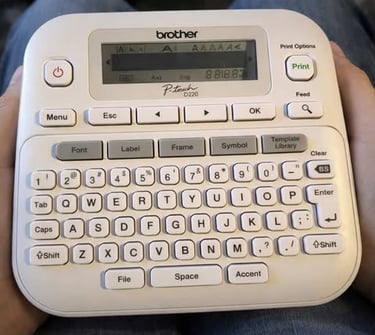 Handheld Brother P-touch label maker with full keyboard and LCD screen resting on a person’s lap