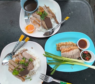 Plates of authentic Thai roasted duck over rice and crispy pork belly served with dipping sauces on a table.