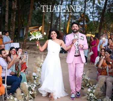 A happy bride in a white gown and groom in a pink suit celebrate their beach wedding in Thailand.