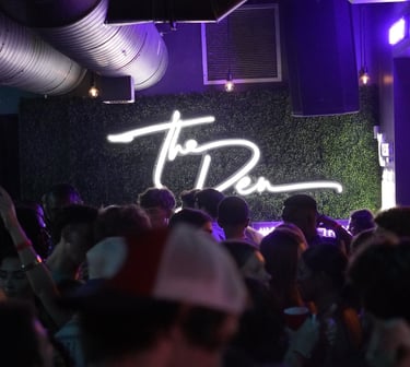 Crowd dancing at The Den nightclub under a white neon sign on a green foliage wall with purple lighting.