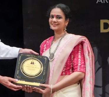 a woman in a white and pink saree receiving an award. She is Ms. Anushree Ladia, Bharatnatyam Expert