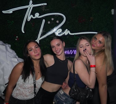 Four young women posing together at a nightclub under a white neon The Den sign.