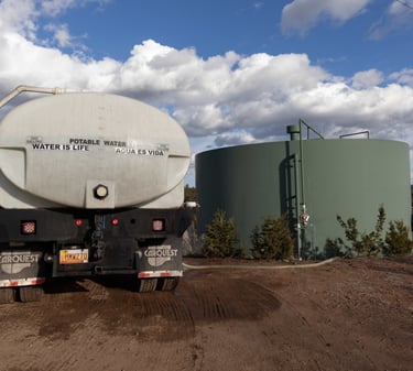 A potable water truck delivery service filling a large green storage tank on a rural landscape.