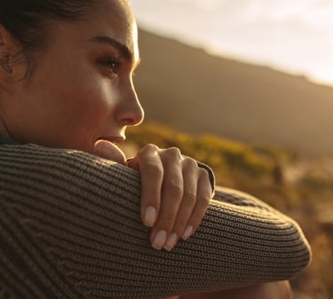 A young female hiker in a grey sweater looks at the golden sunset over mountain hills.