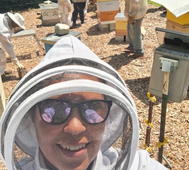 A smiling beekeeper in a protective veil takes a selfie with a group tending to beehives.
