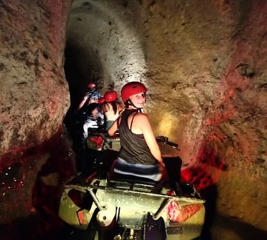 Adventurous tourists wearing red helmets ride ATVs through a narrow limestone underground cave tunnel.