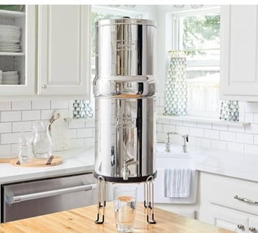 Berkey stainless steel countertop gravity water filter system on a wooden stand in a bright kitchen.