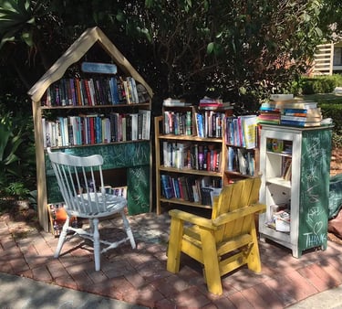 Outdoor community library on a brick patio featuring wooden bookshelves, a white wooden chair, and a yellow chair.