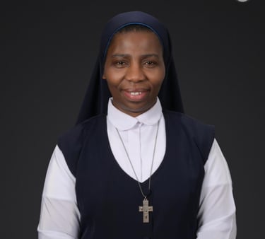 A smiling Catholic nun wearing a blue and white habit with a silver crucifix necklace against a dark background.
