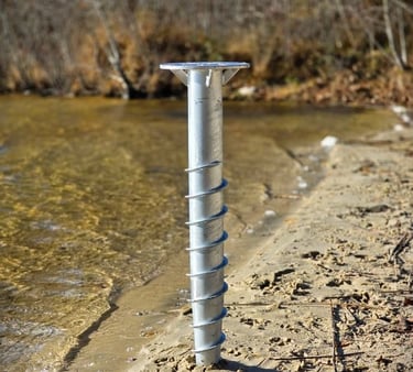 A galvanized steel helical ground screw anchor installed in the sandy shoreline of a lake for dock construction.