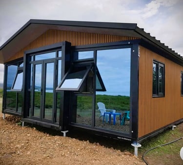 Modern prefab cabin with wood siding and large windows reflecting a green landscape.
