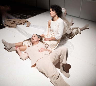 Eine Theaterszene. Drei hell gekleidete Frauen sitzen einträchtig auf dem Boden.