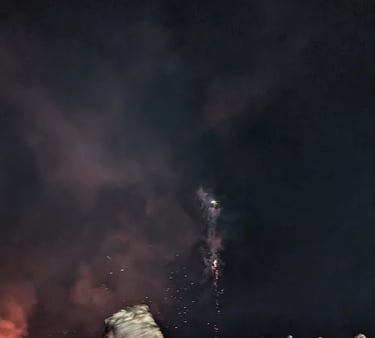 A large crowd of people gathered at night watching a fireworks display and bonfire with smoke in the sky.