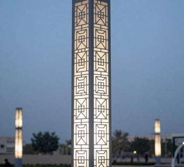 A close-up photograph of a beautifully crafted decorative lighting pole with intricate geometric details, installed in a modern Middle Eastern / Gulf urban park during the blue hour, featuring soft Slate Blue and Off-White tones.