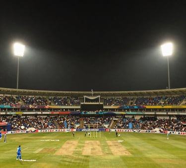 A wide landscape shot of a packed cricket stadium in India at night, vibrant atmosphere with massive floodlights creating a high-energy glow against a dark sky, amber and dark navy color tones.