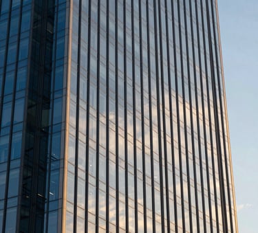 A high-angle photograph of a modern glass skyscraper reflecting a clear sky, with the deep blue glass panels contrasting against the warm golden glow of late afternoon sunlight.