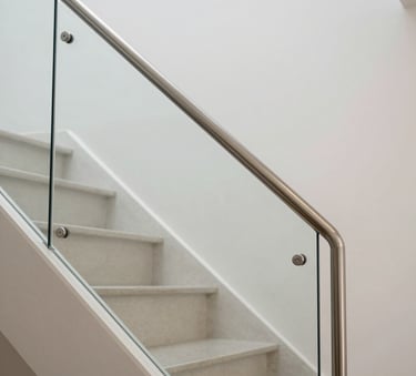 A close-up photograph of a minimalist staircase featuring a polished stainless steel handrail and clear glass safety panels in a modern Hispanic / Latin American luxury home, bright natural lighting.