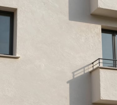A close-up photograph of a high-quality textured facade finish on a Southern European building. The surface is a clean off-white with a modern matte texture, reflecting soft morning sunlight. The composition is architectural and precise, highlighting the professional application of exterior insulation materials.