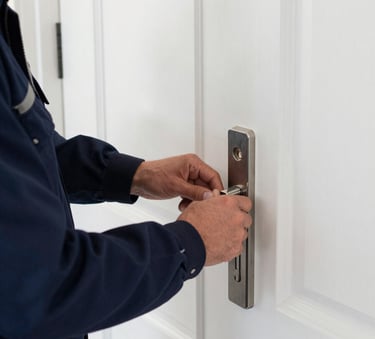 A professional locksmith in a dark blue uniform inspecting a high-security silver door lock on a white front door in a clean Northern European / British residential setting.
