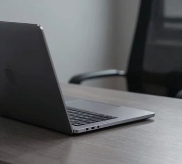A close-up photograph of a sleek, dark charcoal laptop sitting on a polished shadow grey desk in a minimalist corporate office. The lighting is sharp and directional, highlighting the clean lines and authoritative atmosphere of a modern tech hub.