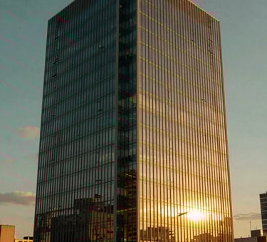 A modern commercial building with glass facades in a South American / Brazilian city at sunset. The lighting is warm and golden, reflecting off the windows, with a sky in deep forest green and soft blue tones.