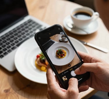 A professional overhead shot of hands using a smartphone to photograph a beautifully plated dish in a sunlit European restaurant, with a laptop and a cup of coffee nearby, warm lighting, elegant and modern style.