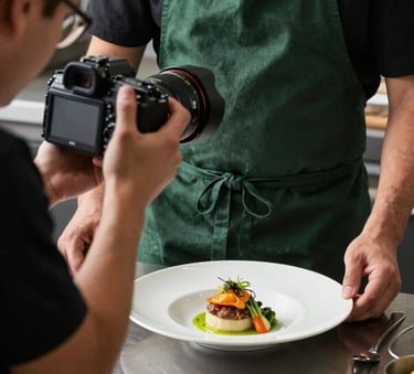A behind-the-scenes shot in a professional North American kitchen setting. A content creator is using a high-end camera to photograph a chef plating a dish. The chef wears a matte forest green apron. The lighting is sophisticated and natural, highlighting the textures of the food.