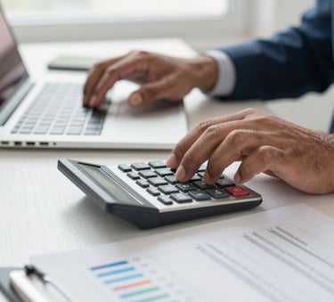 A close-up photograph of a professional's hands in a brightly lit South Asian office, using a calculator alongside organized financial documents and a laptop, reflecting a clean and modern workspace.