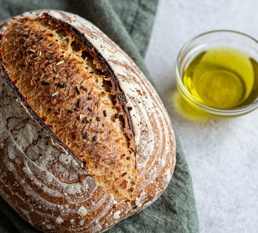 A close-up shot of an artisanal sourdough loaf on a matte forest green linen cloth, next to a small bowl of olive oil on a crisp parchment surface. Soft, Scandinavian-style natural light.
