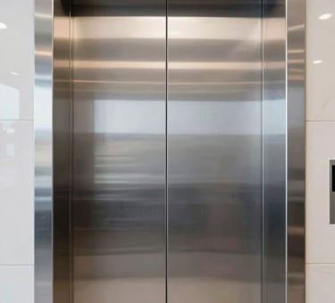 A close-up photograph of sleek, polished silver stainless steel elevator doors in a brightly lit, modern corporate lobby in a South American / Colombian city, professional and clean aesthetic.